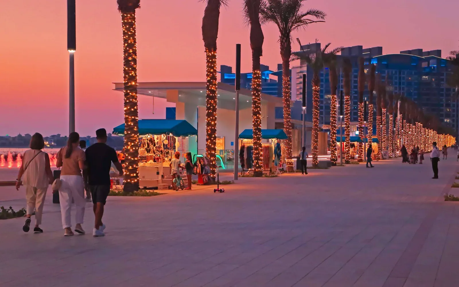 Visitors walking beside illuminated palm trees and kiosks on Palm West Beach promenade at sunset